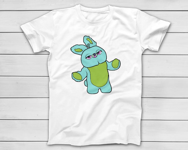 Puede incluir: Camiseta blanca con una ilustraci&oacute;n de dibujos animados de un personaje de conejo azul y verde con ojos rosados. El conejo tiene los brazos extendidos. El dise&ntilde;o est&aacute; centrado en la parte delantera de la camiseta.