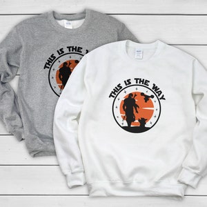 Puede incluir: Dos sudaderas, una gris y otra blanca, con un dise&ntilde;o gr&aacute;fico circular con el texto "THIS IS THE WAY". El dise&ntilde;o incluye una silueta de un personaje, una criatura y una nave espacial sobre un fondo naranja.