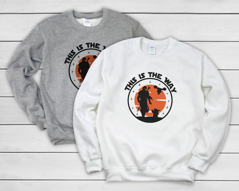 Puede incluir: Dos sudaderas, una gris y otra blanca, con un dise&ntilde;o gr&aacute;fico circular con el texto "THIS IS THE WAY". El dise&ntilde;o incluye una silueta de un personaje, una criatura y una nave espacial sobre un fondo naranja.