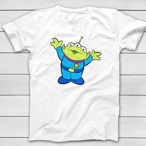 Puede incluir: Camiseta blanca con un personaje alien&iacute;gena de dibujos animados. El alien&iacute;gena tiene tres ojos, piel verde y un atuendo azul y morado. El alien&iacute;gena est&aacute; sonriendo y tiene los brazos levantados. El dise&ntilde;o est&aacute; centrado en la camiseta.