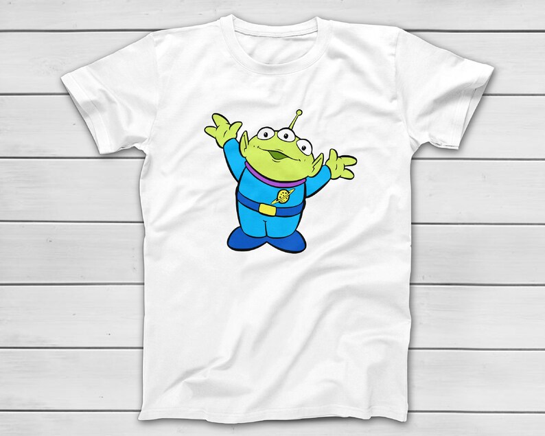 Puede incluir: Camiseta blanca con un personaje alien&iacute;gena de dibujos animados. El alien&iacute;gena tiene tres ojos, piel verde y un atuendo azul y morado. El alien&iacute;gena est&aacute; sonriendo y tiene los brazos levantados. El dise&ntilde;o est&aacute; centrado en la camiseta.