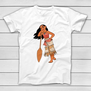 Puede incluir: Camiseta blanca con una ilustraci&oacute;n de dibujos animados de una mujer con un atuendo de inspiraci&oacute;n nativa americana. El personaje est&aacute; apoyado en una paleta de madera. La mujer lleva un top color coral, una falda con flecos y un collar.