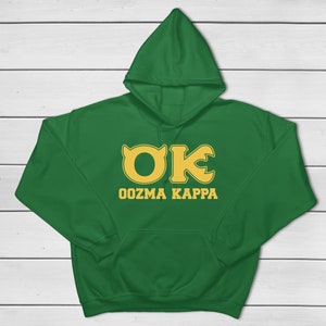 Peut inclure: Sweat-shirt à capuche vert avec une poche kangourou et un cordon de serrage. Le sweat-shirt porte l'inscription "OK Oozma Kappa" en jaune, un design inspiré d'une fraternité universitaire fictive.