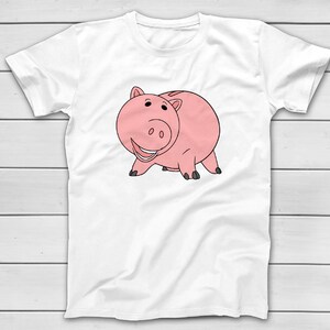 Puede incluir: Camiseta blanca con una ilustraci&oacute;n de dibujos animados de un cerdo rosa con contornos negros. El cerdo est&aacute; sonriendo y de pie sobre sus cuatro patas. El dise&ntilde;o est&aacute; centrado en la parte delantera de la camiseta.