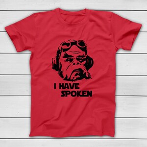 May include: A red t-shirt featuring a black graphic of a character wearing goggles and a headset. Below the graphic, the text "I HAVE SPOKEN" is printed in a bold, sans-serif font. The shirt is displayed on a white wooden surface.