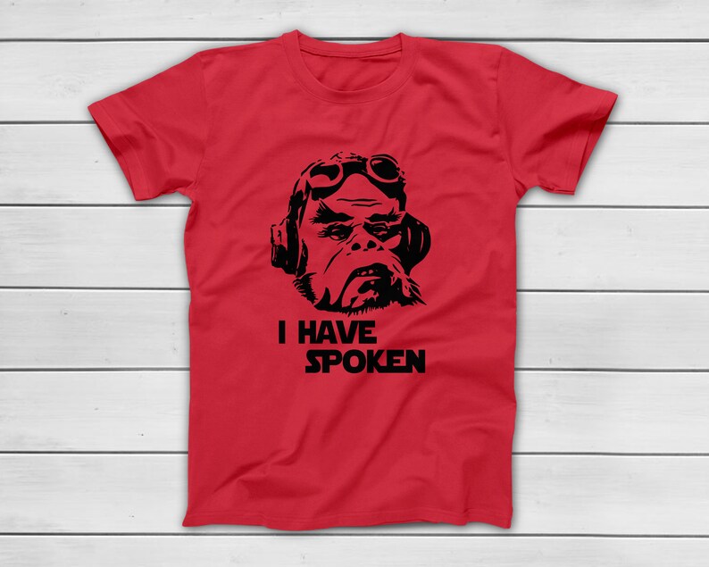 May include: A red t-shirt featuring a black graphic of a character wearing goggles and a headset. Below the graphic, the text "I HAVE SPOKEN" is printed in a bold, sans-serif font. The shirt is displayed on a white wooden surface.