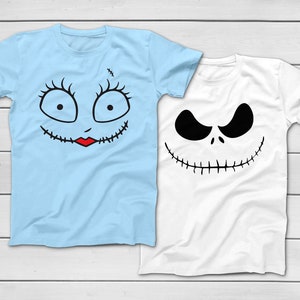 May include: Two short-sleeved t-shirts. One is light blue with a stitched face design, including large eyes, eyelashes, and a red mouth. The other is white with a black face design, featuring a wide grin and angry eyes. Both shirts are on a white wooden surface.