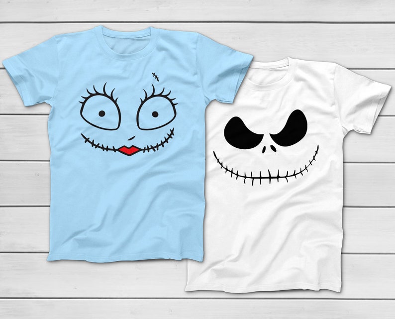 May include: Two short-sleeved t-shirts. One is light blue with a stitched face design, including large eyes, eyelashes, and a red mouth. The other is white with a black face design, featuring a wide grin and angry eyes. Both shirts are on a white wooden surface.
