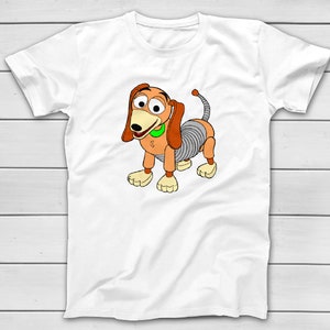Puede incluir: Camiseta blanca con una ilustraci&oacute;n de dibujos animados de Slinky Dog de Toy Story. El perro est&aacute; representado en tonos marrones, beige y grises, con un collar verde. El dise&ntilde;o est&aacute; centrado en la camiseta.