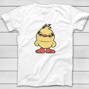 Puede incluir: Camiseta blanca con un dise&ntilde;o de pollito de dibujos animados. El pollito tiene un cuerpo amarillo, pies rojos y maquillaje de ojos morado, con una expresi&oacute;n ligeramente gru&ntilde;ona. La camiseta se muestra sobre una superficie de madera blanca.