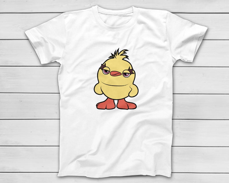 Puede incluir: Camiseta blanca con un dise&ntilde;o de pollito de dibujos animados. El pollito tiene un cuerpo amarillo, pies rojos y maquillaje de ojos morado, con una expresi&oacute;n ligeramente gru&ntilde;ona. La camiseta se muestra sobre una superficie de madera blanca.