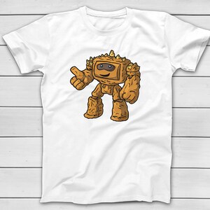 Puede incluir: Camiseta blanca con un personaje de dibujos animados que se asemeja a un golem de roca. El personaje marr&oacute;n sonr&iacute;e y hace un gesto de pulgar hacia arriba. El dise&ntilde;o est&aacute; centrado en la camiseta.