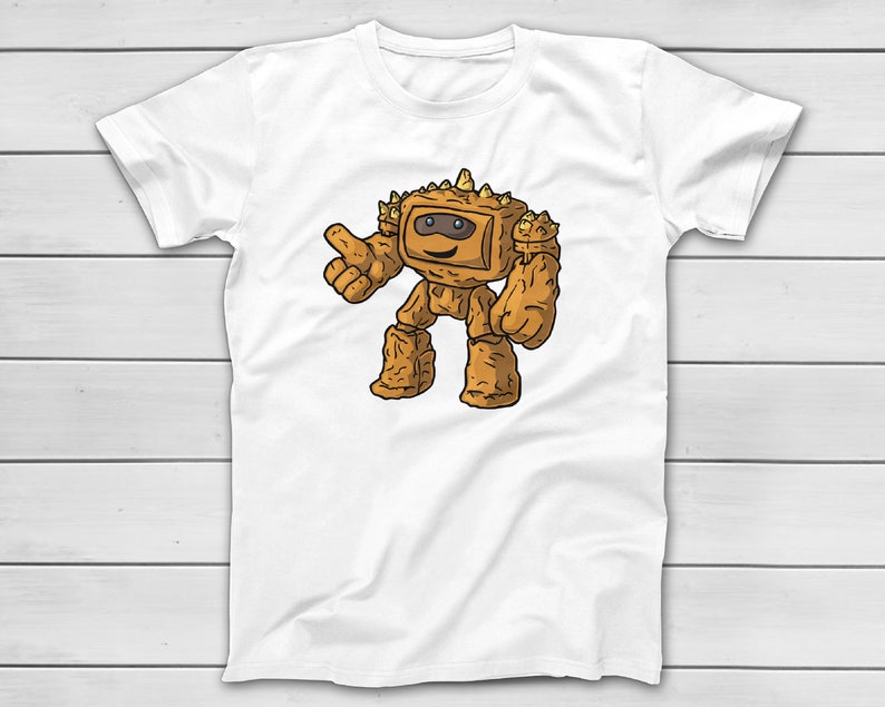 Puede incluir: Camiseta blanca con un personaje de dibujos animados que se asemeja a un golem de roca. El personaje marr&oacute;n sonr&iacute;e y hace un gesto de pulgar hacia arriba. El dise&ntilde;o est&aacute; centrado en la camiseta.