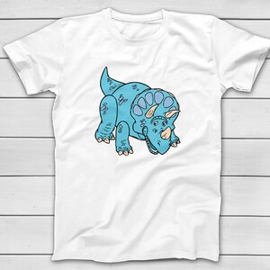 Puede incluir: Camiseta blanca con una ilustraci&oacute;n de dibujos animados de un dinosaurio Triceratops azul. El dinosaurio tiene un cuerpo azul claro, un volante y tres cuernos. El dise&ntilde;o est&aacute; centrado en la parte delantera de la camiseta.