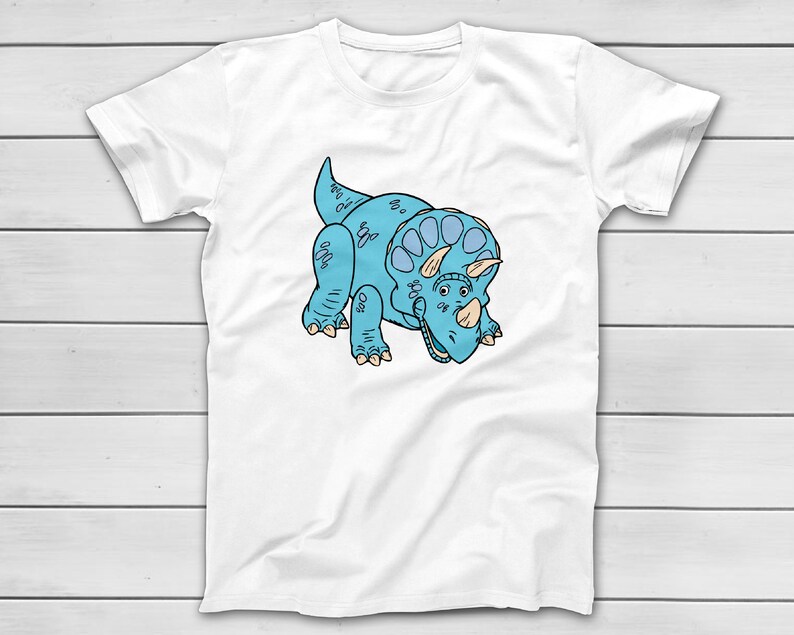 Puede incluir: Camiseta blanca con una ilustraci&oacute;n de dibujos animados de un dinosaurio Triceratops azul. El dinosaurio tiene un cuerpo azul claro, un volante y tres cuernos. El dise&ntilde;o est&aacute; centrado en la parte delantera de la camiseta.