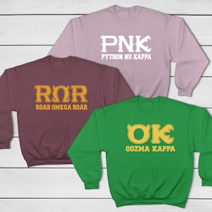 Peut inclure: Trois sweat-shirts à col ras du cou, de couleur bordeaux, violet clair et vert. Chaque sweat-shirt présente un design différent avec des lettres dorées. Le sweat-shirt bordeaux porte l'inscription "ROR ROAR OMEGA ROAR". Le sweat-shirt violet clair porte l'inscription "PNK PYTHON NU KAPPA". Le sweat-shirt vert porte l'inscription "OK OOZMA KAPPA".