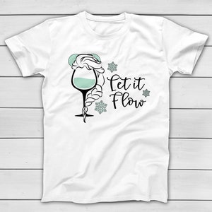 May include: White t-shirt featuring a whimsical design of a wine glass with a braided hairstyle and the text "Let it Flow" in black script. The glass is filled with a light teal liquid, and snowflakes surround the text.