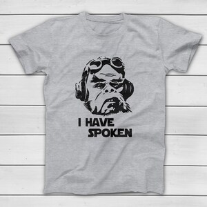 May include: A light grey t-shirt with a black graphic of a character wearing goggles and headphones. The text "I HAVE SPOKEN" is printed below the image. The shirt is made of a soft material.