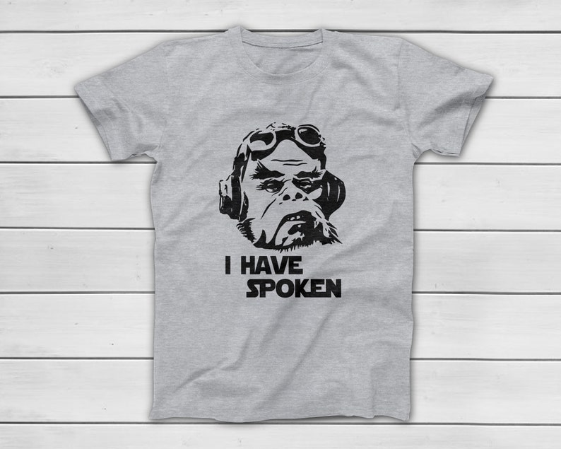 May include: A light grey t-shirt with a black graphic of a character wearing goggles and headphones. The text "I HAVE SPOKEN" is printed below the image. The shirt is made of a soft material.