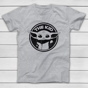 May include: A light grey t-shirt with a black and white graphic of a character from a popular science fiction series. The graphic is a circular design with the text "THE KID" around the character's image.