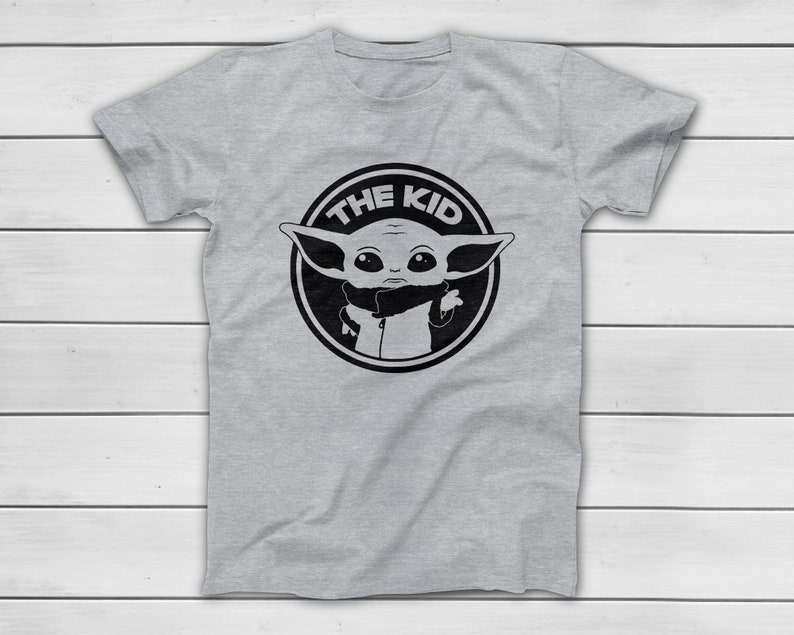 May include: A light grey t-shirt with a black and white graphic of a character from a popular science fiction series. The graphic is a circular design with the text "THE KID" around the character's image.