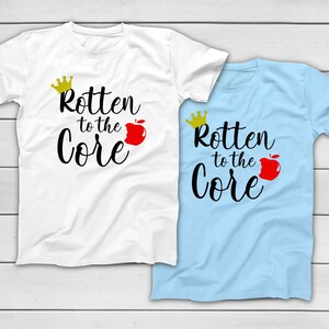 May include: Two t-shirts, one white and one light blue, each with the phrase "Rotten to the Core" in black script. A gold crown and a red apple graphic accompany the text, creating a playful design. The shirts are likely made of cotton.