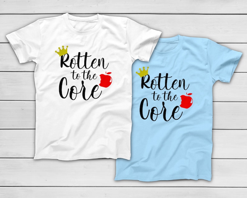 May include: Two t-shirts, one white and one light blue, each with the phrase "Rotten to the Core" in black script. A gold crown and a red apple graphic accompany the text, creating a playful design. The shirts are likely made of cotton.