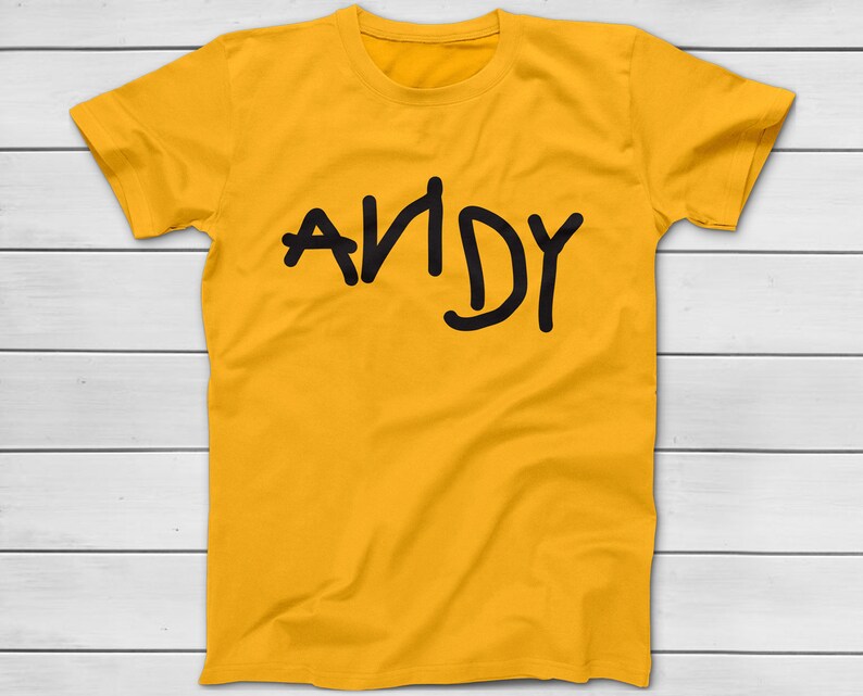 Puede incluir: Camiseta de color amarillo mostaza con el nombre "ANDY" impreso en negro, con letras manuscritas. La camiseta est&aacute; hecha de un material suave y tiene mangas cortas. El texto est&aacute; ligeramente descentrado.