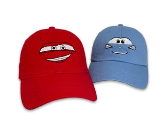 May include: Two baseball caps, one red and one light blue, each embroidered with a cartoon car face. The red cap has a smiling face design, while the blue cap has a different car face design.