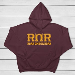 Peut inclure: Un sweat à capuche bordeaux avec une poche avant. Le devant affiche le texte "ROR ROAR OMEGA ROAR" dans une police dorée stylisée. Le sweat-shirt est présenté sur un fond en bois blanc.