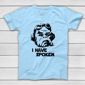 May include: Pale blue t-shirt with a black and white graphic of a character wearing goggles and headphones. The text "I HAVE SPOKEN" is printed below the graphic in a bold, black font. The shirt is displayed on a white wooden surface.