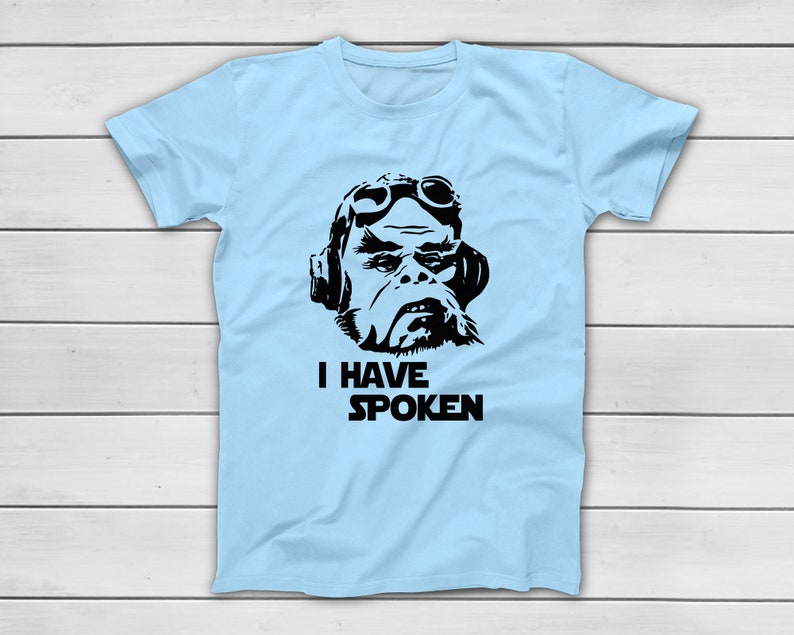 May include: Pale blue t-shirt with a black and white graphic of a character wearing goggles and headphones. The text "I HAVE SPOKEN" is printed below the graphic in a bold, black font. The shirt is displayed on a white wooden surface.