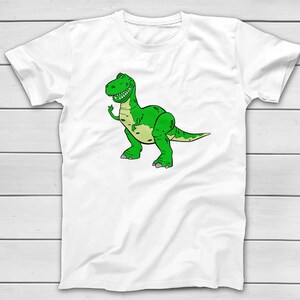 Puede incluir: Camiseta blanca con una ilustraci&oacute;n de dibujos animados de un Tyrannosaurus Rex verde. El dinosaurio tiene una expresi&oacute;n amigable y un vientre verde claro. El dise&ntilde;o est&aacute; sobre un fondo blanco.