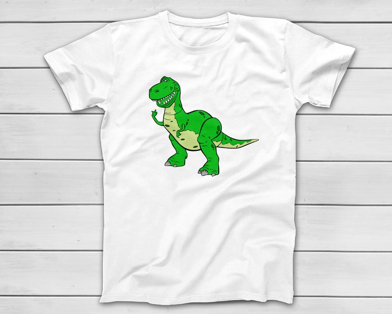 Puede incluir: Camiseta blanca con una ilustraci&oacute;n de dibujos animados de un Tyrannosaurus Rex verde. El dinosaurio tiene una expresi&oacute;n amigable y un vientre verde claro. El dise&ntilde;o est&aacute; sobre un fondo blanco.