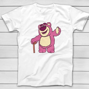 Puede incluir: Camiseta blanca con una ilustraci&oacute;n de dibujos animados de un oso rosa con un bast&oacute;n marr&oacute;n. El oso est&aacute; sonriendo y saludando. El dise&ntilde;o est&aacute; impreso en la parte delantera de la camiseta.