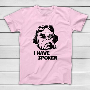 May include: Pale pink t-shirt featuring a black graphic of a character wearing goggles and a headset. The text "I HAVE SPOKEN" is printed below the character's face. The shirt has short sleeves and a crew neck.