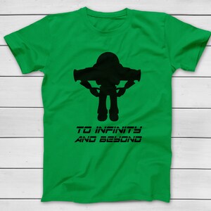 May include: A green t-shirt featuring a black silhouette of a space ranger with outstretched arms. Below the image is the text "TO INFINITY AND BEYOND" in a stylized font. The shirt is displayed on a white wooden surface.