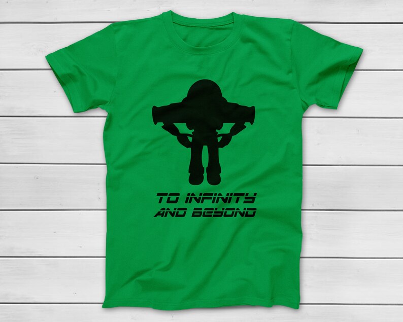 May include: A green t-shirt featuring a black silhouette of a space ranger with outstretched arms. Below the image is the text "TO INFINITY AND BEYOND" in a stylized font. The shirt is displayed on a white wooden surface.