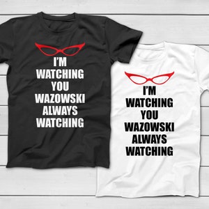 Peut inclure: Deux t-shirts, un noir et un blanc, avec le même graphisme. Le graphisme comprend des lunettes rouges en œil de chat au-dessus du texte "I'M WATCHING YOU WAZOWSKI ALWAYS WATCHING" en gras, police sans-serif.