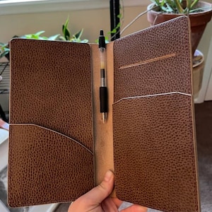 May include: Open brown leather journal with a pen in the centre. The journal has pockets on the left and right sides. The pen is black with a clear barrel. The journal is open, revealing the interior.