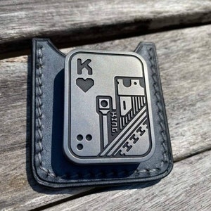 May include: A silver metal card holder with a black leather pocket. The card holder features a black and white design of a playing card with the King of Hearts and a building.