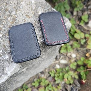 May include: Two black leather keychains with stitched edges. One keychain has red stitching, the other has black stitching. The keychains are rectangular and appear to be handmade.