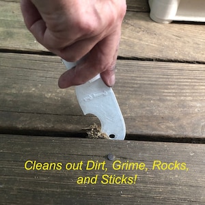 May include: A white plastic tool with a curved edge and a hole is being used to clean out dirt, grime, rocks, and sticks from between wooden deck boards. The text "Cleans out Dirt, Grime, Rocks, and Sticks!" is written in yellow on the wooden surface.