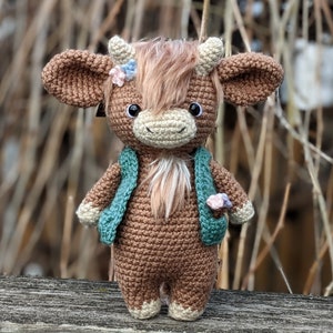 May include: A brown crocheted stuffed animal cow with a green waistcoat and pink flowers. The cow has a fluffy brown mane and a white face.