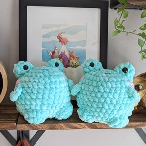 Op de afbeelding: Twee gehaakte tealblauwe kikker knuffels met zwarte ogen en roze accenten die op een houten plank staan.