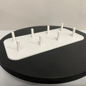 May include: White plastic sewing needle holder with eight small white pegs. The holder is sitting on a black circular base.