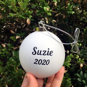 Pode incluir: Enfeite de Natal branco com o nome "Suzie" e o ano "2020" escritos em caligrafia preta.