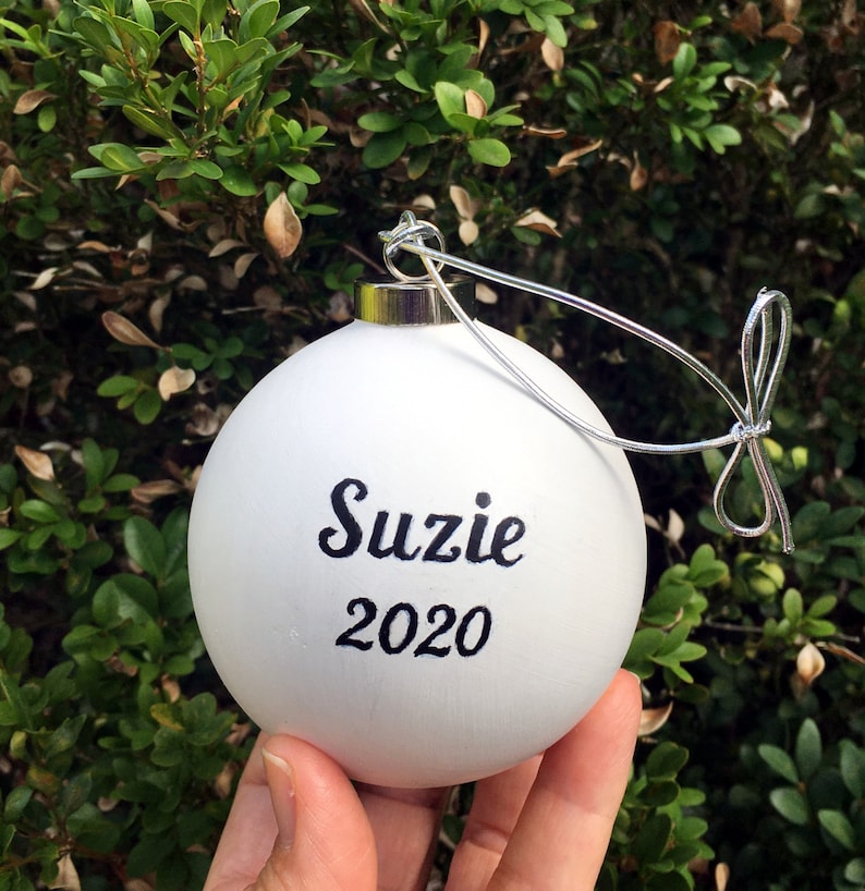 Pode incluir: Enfeite de Natal branco com o nome "Suzie" e o ano "2020" escritos em caligrafia preta.