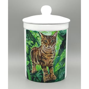 May include: A white ceramic jar with a lid, decorated with a hand-painted image of a brown and black striped cat amidst green leaves. The jar is round with a white knob on top. The jar is approximately 15 cm tall.