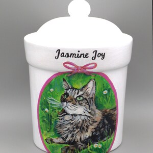 May include: White ceramic container with a lid, featuring a hand-painted portrait of a cat within a green and pink oval. The name "Jasmine Joy" is written above the cat portrait. A pink bow is below the name.